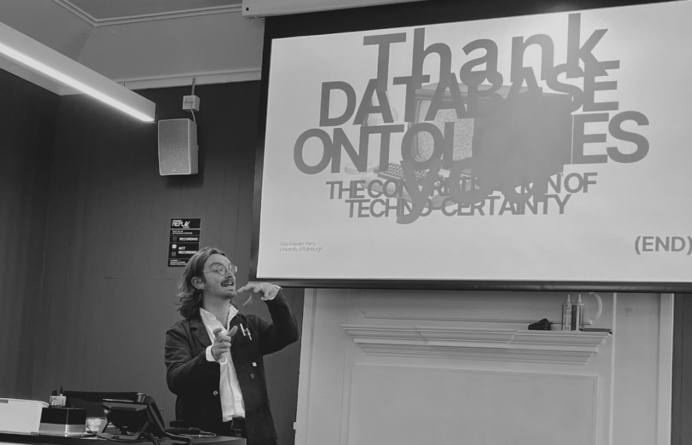 Black and white image of Max presenting at 'social science of database workshop'. Max is stood, gesturing, in front of a screen with the words 'database ontology' written in large font.